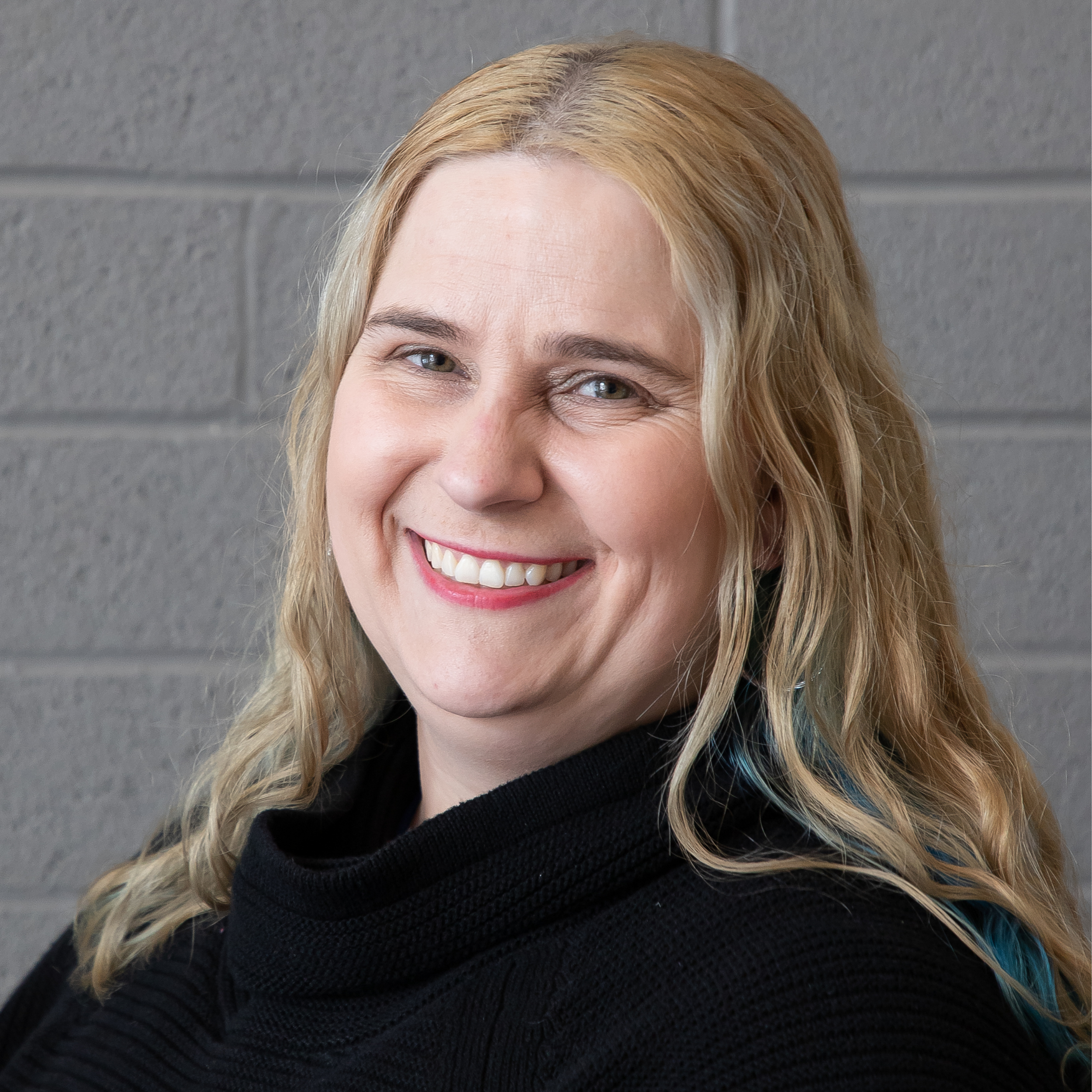 Woman with blonde hair and a black turtleneck smiling in front of a gray brick wall