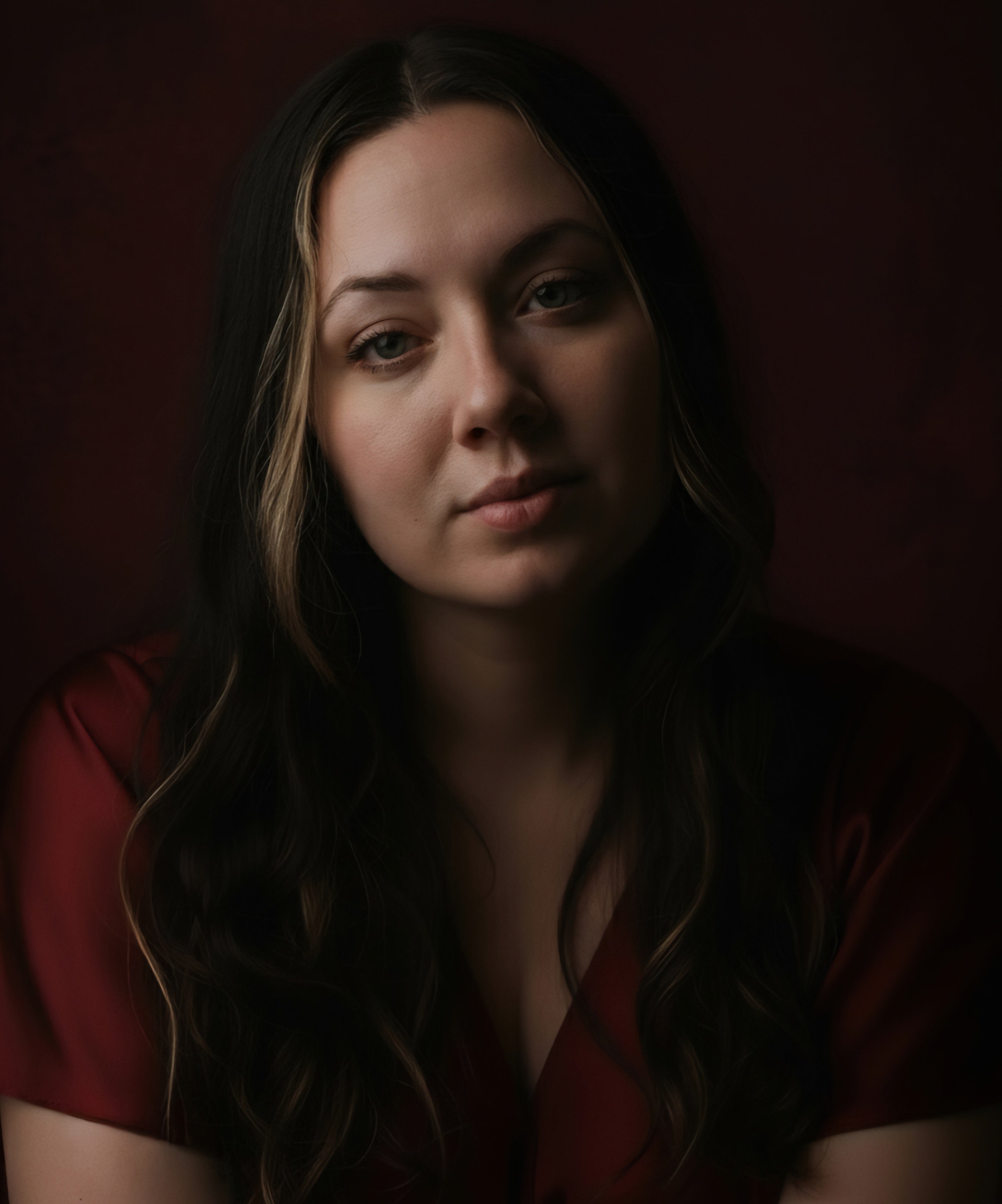Woman with long dark hair wearing a red top against a dark background