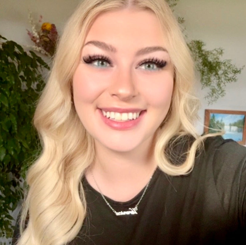 Woman with blonde hair and makeup smiling in front of a plant and framed picture.