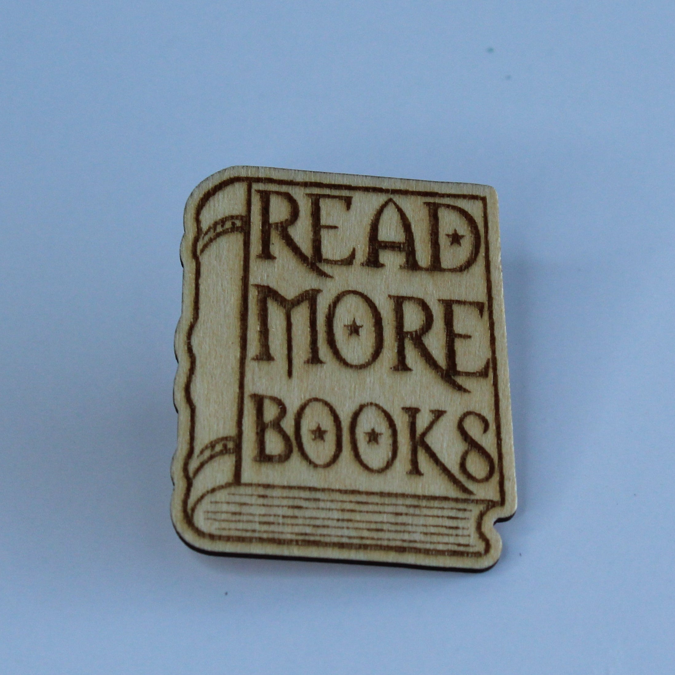 A small book-shaped wooden pin with the words 'READ MORE BOOKS' in dark lettering on a light wood-toned background, centered on a solid light blue backdrop.