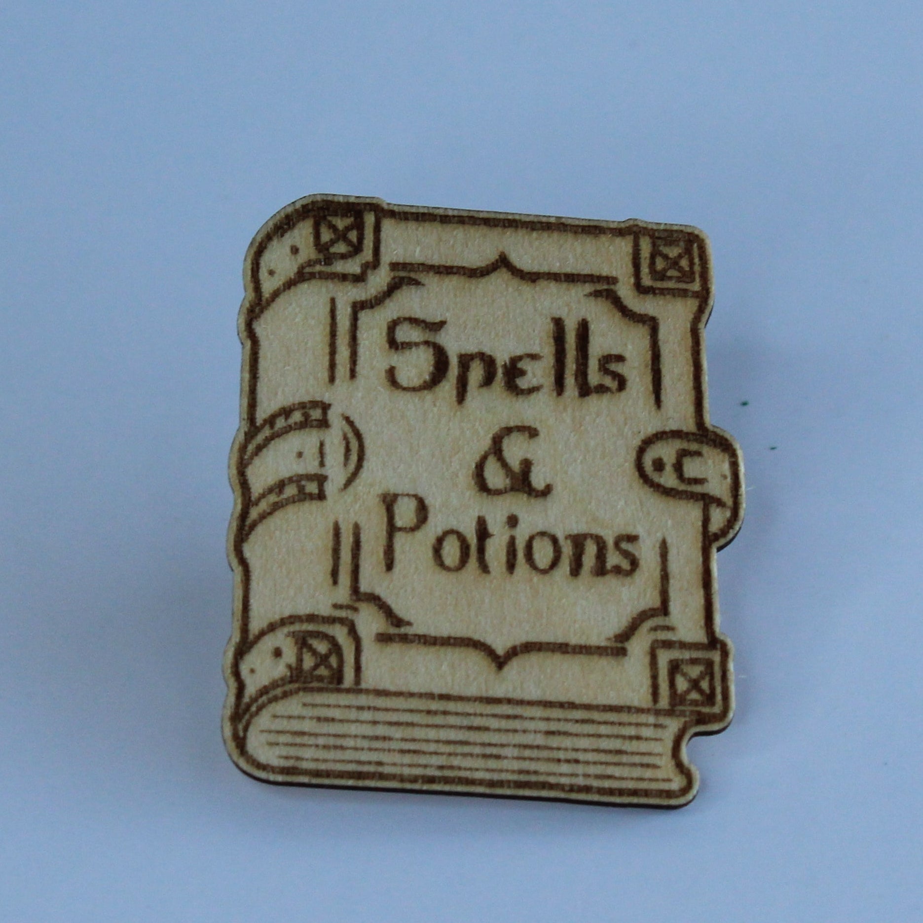 A small book-shaped wooden pin with the words 'Spells & Potions' engraved on the cover, centered against a solid light blue background.