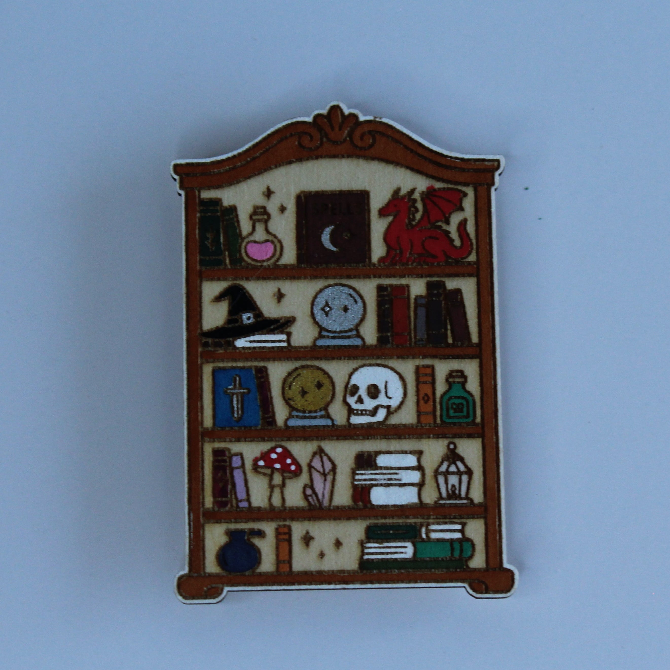 A small enamel pin shaped like a wooden bookshelf filled with various occult and study-themed items, including books, a skull, a potion bottle, a dragon, and a cauldron, set against a solid light blue background.