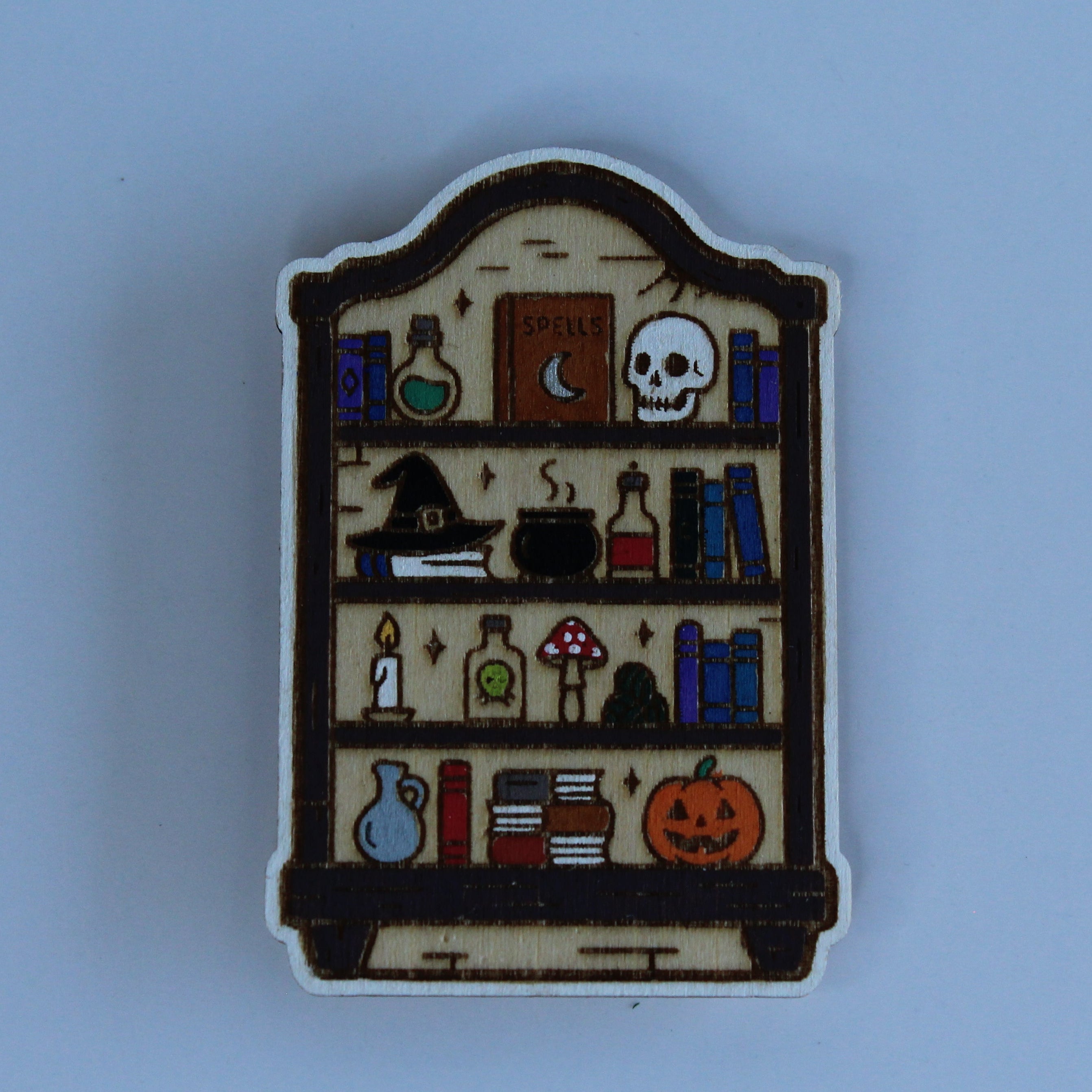 A small arched shelf-shaped pin or badge featuring a Halloween-themed illustration with books, potion bottles, a skull, a cauldron, mushrooms, and a pumpkin on a light blue background.
