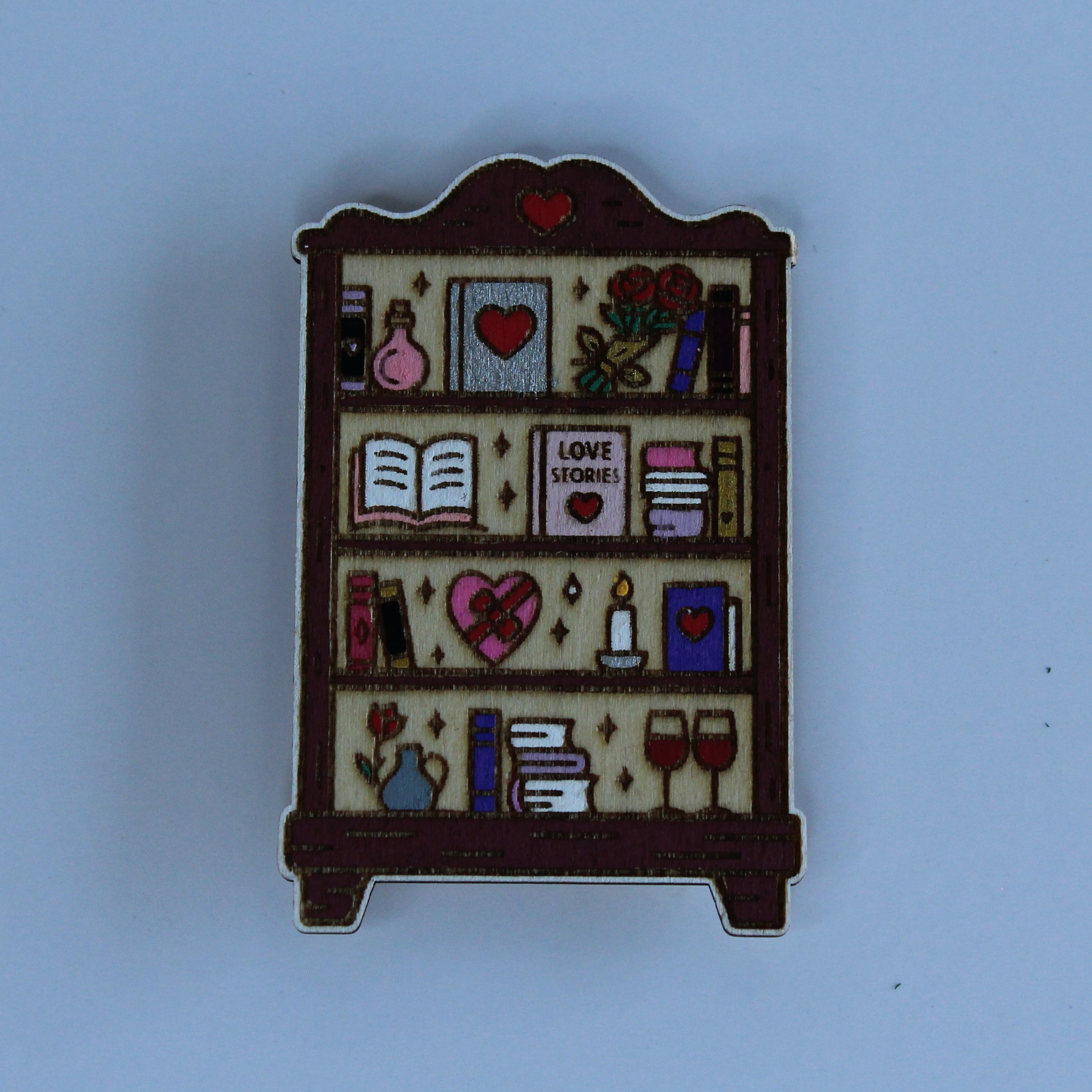 A small enamel pin shaped like a bookshelf filled with miniature books, hearts, flowers, and other decorative items, photographed against a solid light blue background.