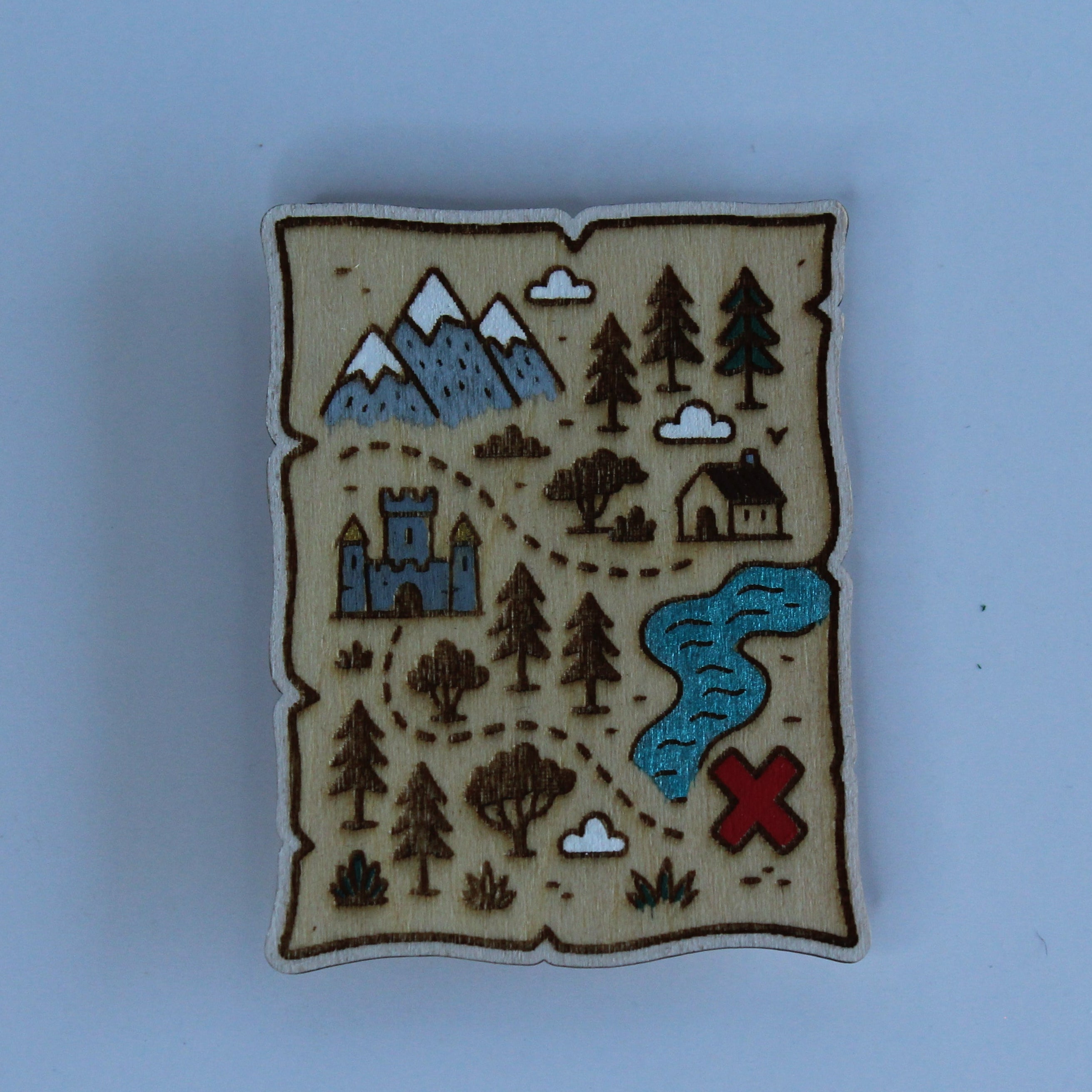 A small rectangular enamel pin shaped like an old treasure map, featuring a beige background with brown borders, blue mountains and river, green trees, and a red X, shown on a light blue background.