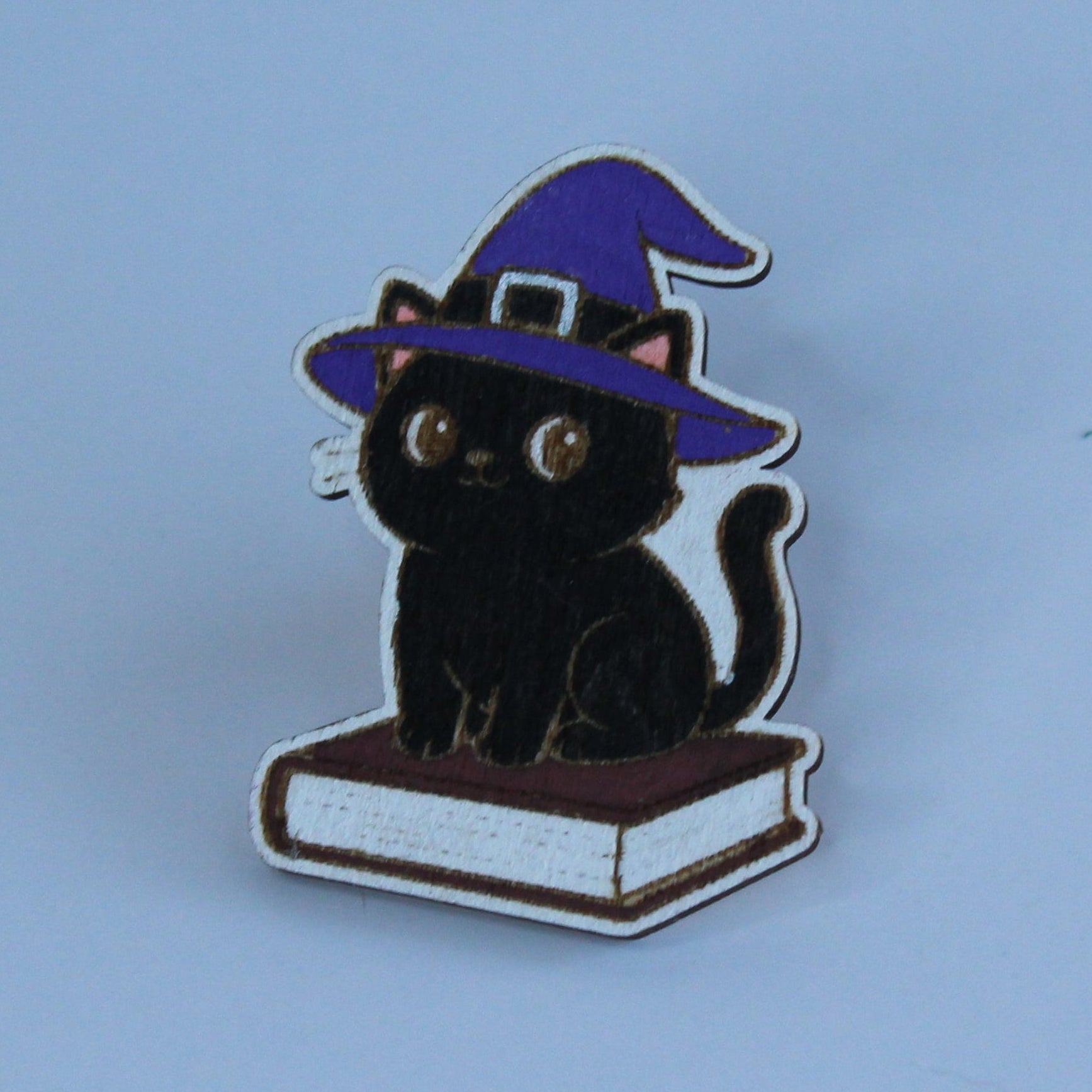 A small wooden pin featuring a black cat wearing a purple witch hat and sitting on a book, photographed against a plain light blue background.