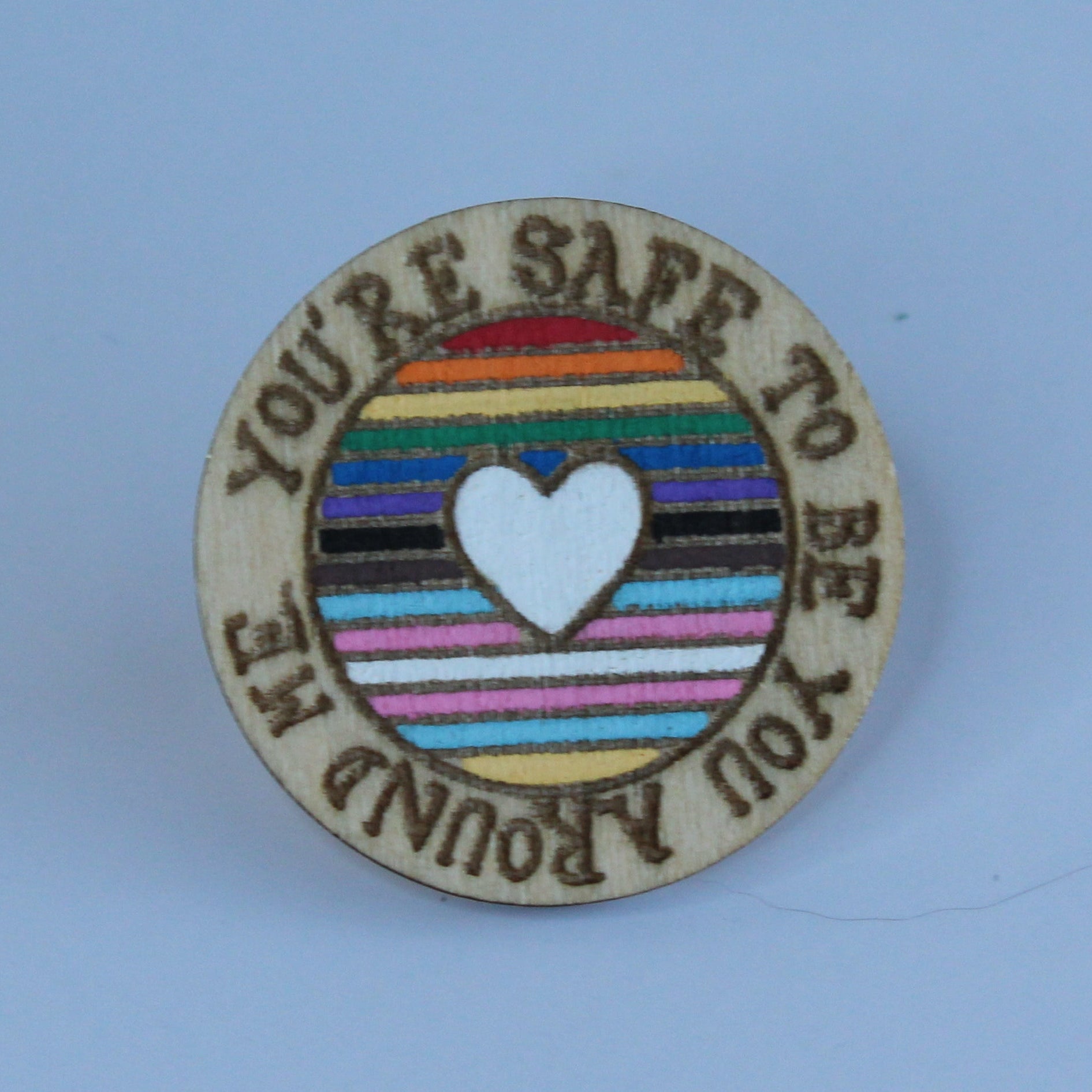 A small circle pin with a rainbow stripe pattern, a white heart in the center, and the text 'YOU'RE SAFE TO BE YOU AROUND YOU' around the edge, shown on a light blue background.