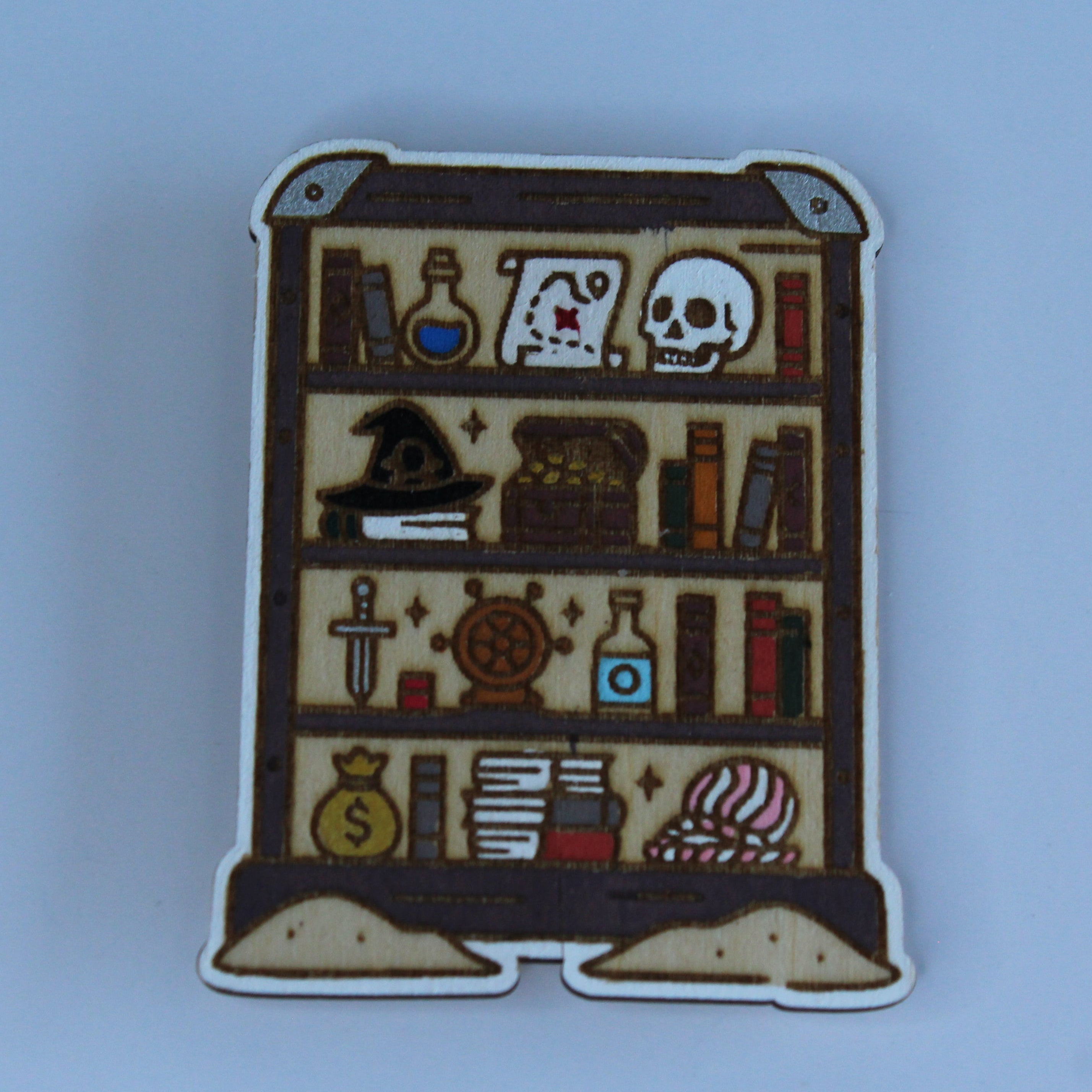 A small enamel pin shaped like a wooden bookshelf filled with occult and fantasy items, including a skull, potion bottles, books, a cauldron, and a wand, shown on a light blue background.