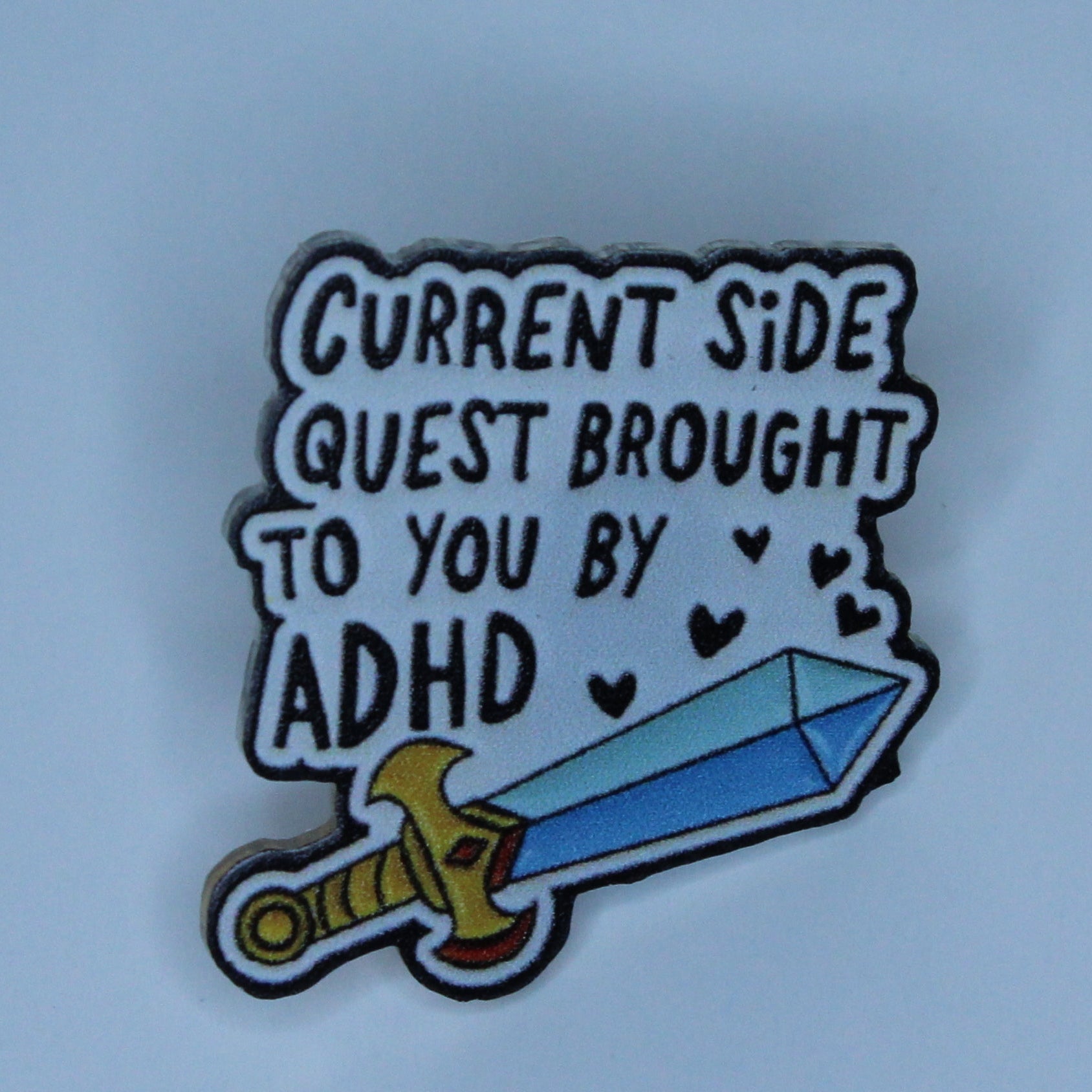 A small acrylic pin on a light blue background. The pin features the text 'CURRENT SIDE QUEST BROUGHT TO YOU BY ADHD' in white lettering with black outlines, above an illustration of a blue sword with a gold hilt.