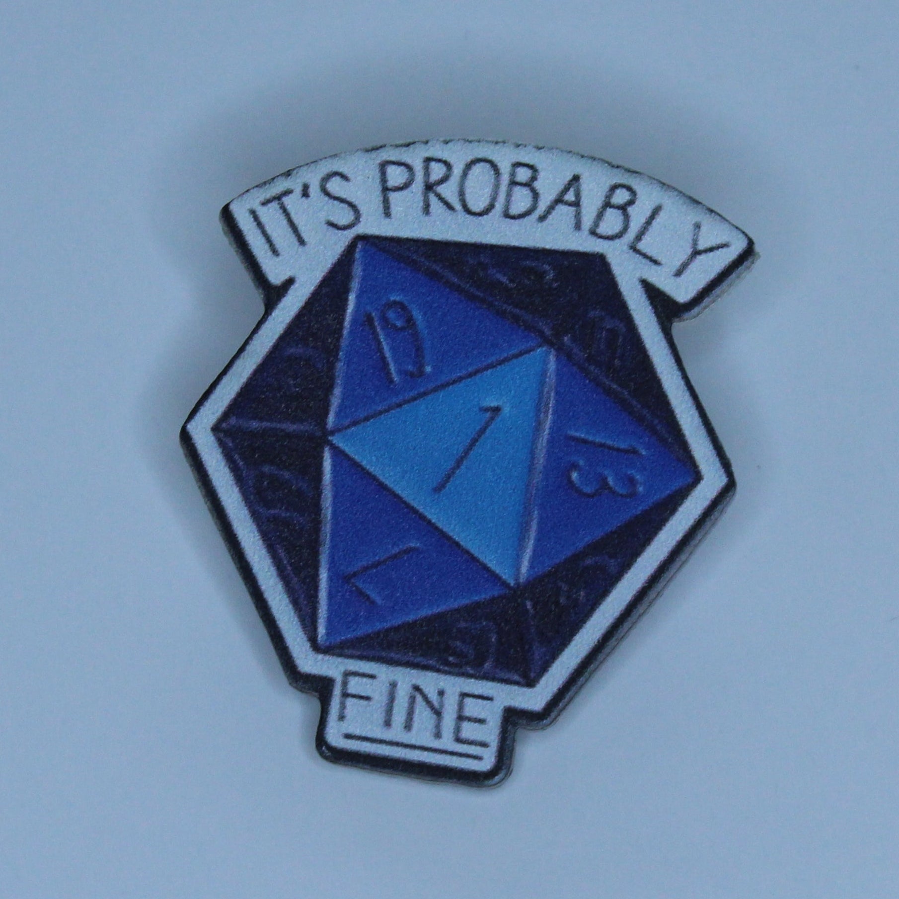 A small acrylic pin shaped like a d20 die, with a blue and dark purple 1, and the text 'IT'S PROBABLY FINE' on a light blue background.