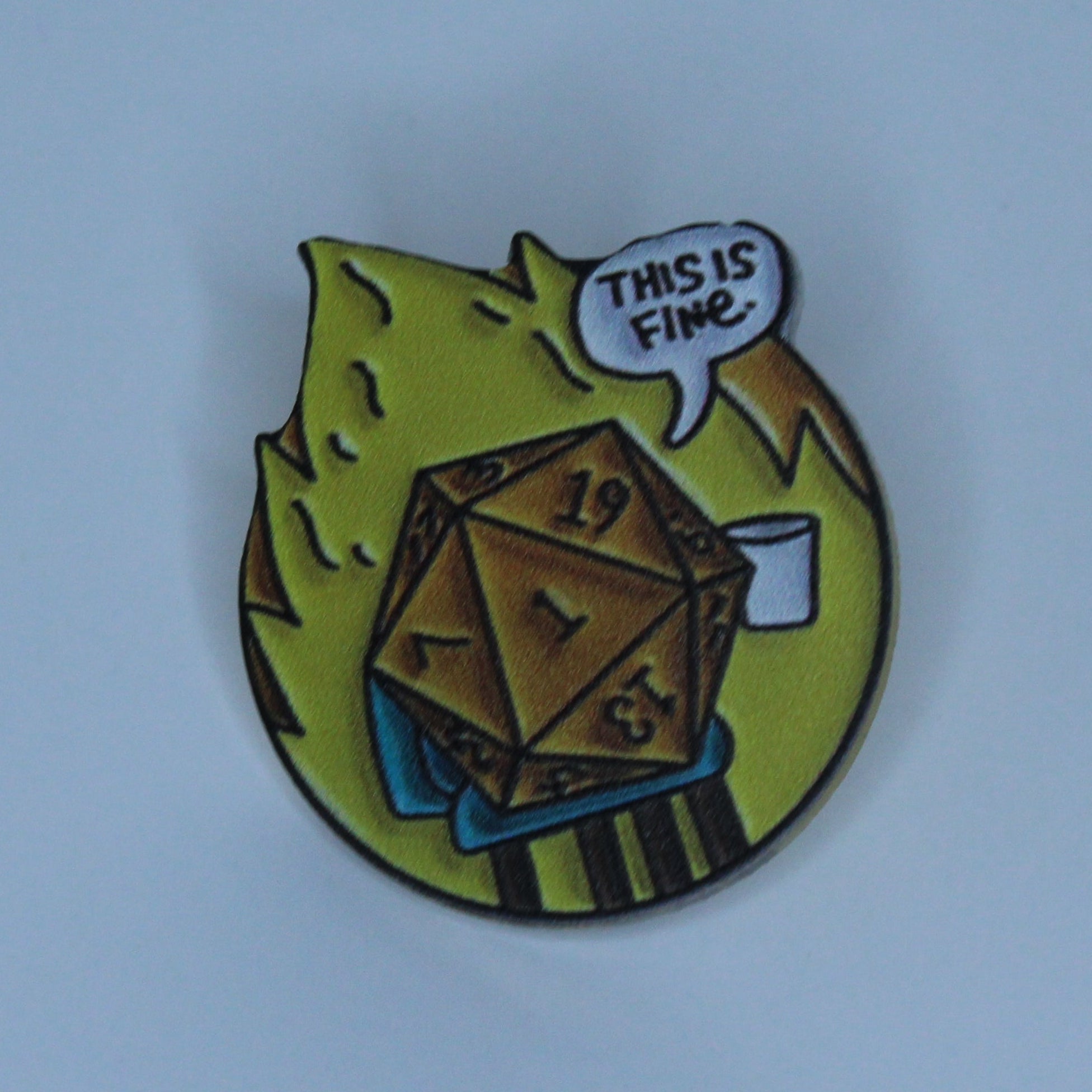 A small acrylic pin featuring a brown 20-sided die rolling on a blue surface with green and yellow flame-like shapes, set against a light blue background.